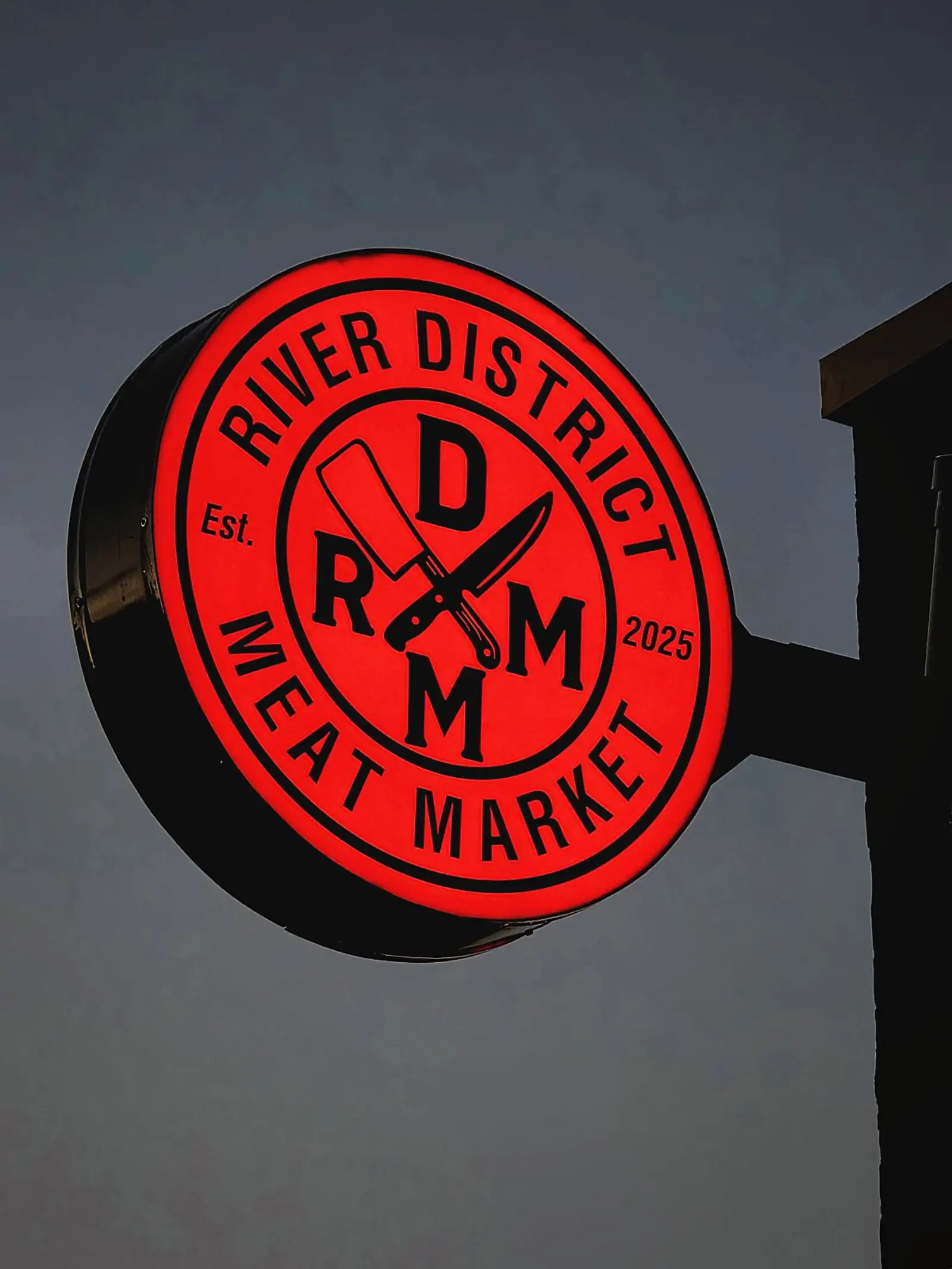 The round River District Meat Market sign glowing red against the evening sky